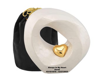 May include: A white, abstract-shaped cremation urn with a gold heart accent. The urn has the text "Always In My Heart David 03/15/1970 - 12/27/2023". A black velvet bag with a gold drawstring is behind the urn.