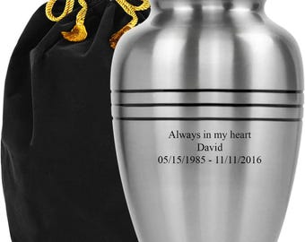 May include: A brushed silver urn with a rounded shape and a lid. The urn has horizontal bands and engraved text that reads "Always in my heart David 05/15/1985 - 11/11/2016." A black velvet bag with a gold drawstring is next to the urn.