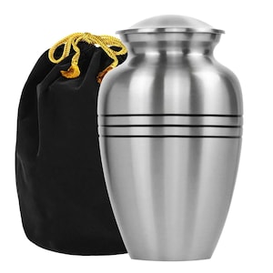 May include: A brushed silver urn with a lid, featuring two horizontal bands. A black velvet bag with a gold drawstring is next to the urn. The urn has a classic, elegant design.