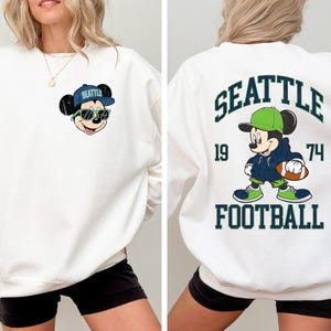 Puede incluir: Sudadera blanca con Mickey Mouse con gorra y gafas de sol de los Seattle Seahawks. La parte trasera de la sudadera muestra "SEATTLE" sobre un dibujo de Mickey Mouse sosteniendo un bal&oacute;n de f&uacute;tbol, con "1974" y "FOOTBALL" debajo.