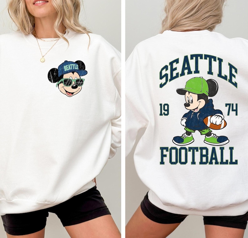 Puede incluir: Sudadera blanca con Mickey Mouse con gorra y gafas de sol de los Seattle Seahawks. La parte trasera de la sudadera muestra "SEATTLE" sobre un dibujo de Mickey Mouse sosteniendo un bal&oacute;n de f&uacute;tbol, con "1974" y "FOOTBALL" debajo.