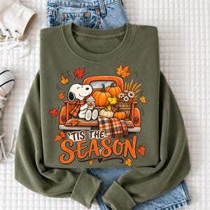 May include: An olive green crewneck sweatshirt featuring a graphic of a white dog sitting in the back of an orange truck filled with pumpkins and wheat. The text "TIS THE SEASON" is printed in orange below the truck, surrounded by autumn leaves.
