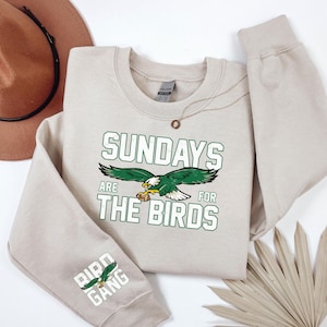 May include: A beige sweatshirt with the text "SUNDAYS ARE FOR THE BIRDS" in green and white, featuring a green and white eagle graphic. The sleeve displays "BIRD GANG" with an eagle. A brown hat and necklace are also visible.
