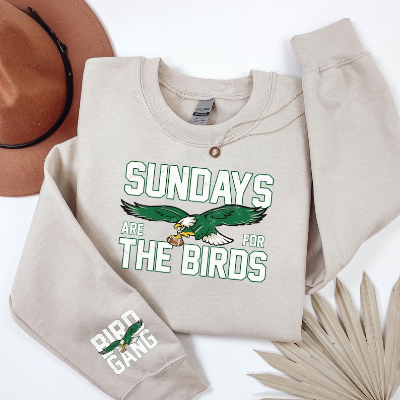 May include: A beige sweatshirt with the text "SUNDAYS ARE FOR THE BIRDS" in green and white, featuring a green and white eagle graphic. The sleeve displays "BIRD GANG" with an eagle. A brown hat and necklace are also visible.