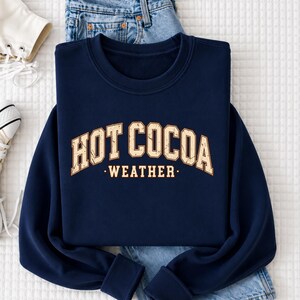 A navy blue sweatshirt with the text "HOT COCOA WEATHER" printed on it, along with a pair of jeans and a white shoe.