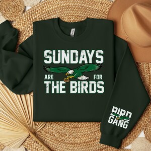 May include: Dark green sweatshirt with the text "SUNDAYS ARE FOR THE BIRDS" in white block letters, featuring a green and white eagle graphic. The right sleeve has "BIRD GANG" in white block letters with a green eagle graphic.