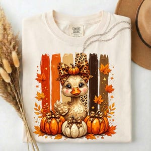 May include: A cream-colored t-shirt featuring a graphic of a duckling wearing a leopard print bow, surrounded by orange and brown pumpkins and autumn leaves. The design includes vertical brushstroke stripes in shades of orange and brown.