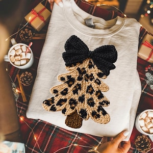 May include: A beige crewneck sweatshirt featuring a central graphic of a stylized Christmas tree with a leopard print pattern and a large black bow at the top. The tree has a brown trunk and a textured, knit-like appearance on the fabric.