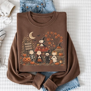 May include: A brown crewneck sweatshirt featuring a graphic print of Peanuts characters in Halloween costumes. The design includes Snoopy, Woodstock, and friends near a doghouse, pumpkins, a crescent moon, and a sign reading PUMPKINS HAY-RIDES CORN MAZE.
