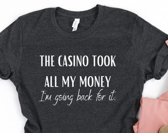 May include: A dark charcoal gray short-sleeve t-shirt with white text that reads THE CASINO TOOK ALL MY MONEY I'm going back for it. The shirt features a crew neckline and rolled sleeves, with a white brand label visible at the inner collar.