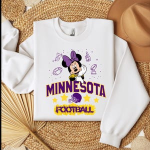 May include: White sweatshirt featuring Minnie Mouse with a purple bow, surrounded by football-themed graphics. The word "MINNESOTA" is displayed in purple and gold, with "FOOTBALL" below. A football helmet and stars complete the design.