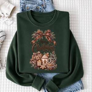 A green sweatshirt with a Christmas-themed design, including a bow, pine cones, and a mug with a candle, is displayed on a white surface.