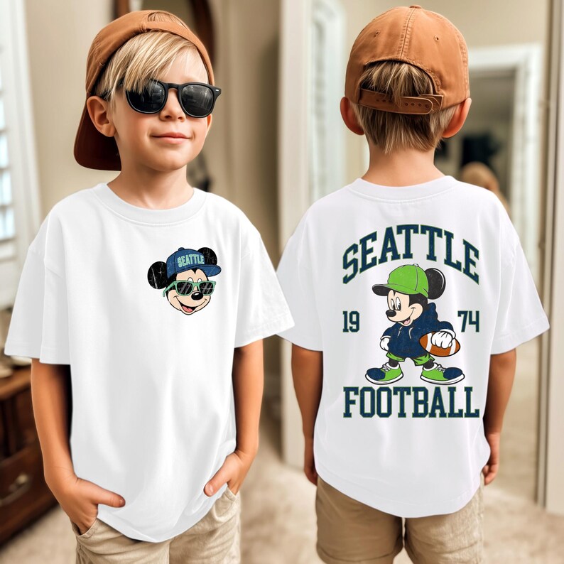 Puede incluir: Camiseta blanca con un dibujo animado de Mickey Mouse con gafas de sol y una gorra azul "Seattle". La parte trasera muestra a Mickey con uniforme de f&uacute;tbol, el texto "SEATTLE FOOTBALL" y los n&uacute;meros "19" y "74".