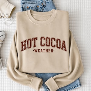 A beige sweatshirt with the text "HOT COCOA WEATHER" printed on it, along with a pair of blue jeans and white sneakers.