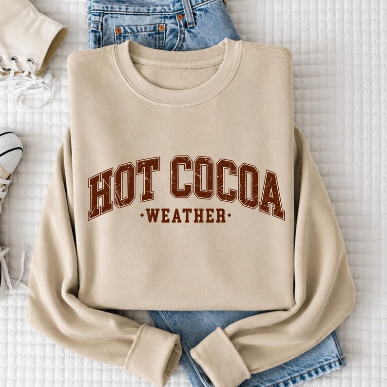 A beige sweatshirt with the text "HOT COCOA WEATHER" printed on it, along with a pair of blue jeans and white sneakers.