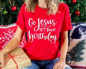 May include: A bright red short-sleeved t-shirt featuring white text that reads Go Jesus IT'S YOUR birthday in a mix of script and sans-serif fonts. The shirt has a crew neckline and is worn with blue denim jeans while holding a gift box.