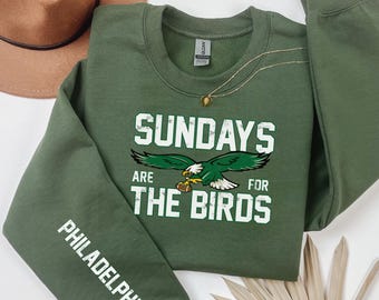 May include: An olive green crewneck sweatshirt featuring white distressed text that reads SUNDAYS ARE FOR THE BIRDS with a graphic of an eagle holding a football. The left sleeve features the white text PHILADELPHIA in a vertical orientation.