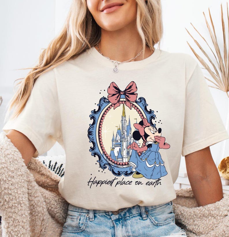 May include: Cream-colored t-shirt featuring a cartoon Minnie Mouse in a blue dress, with a pink bow and a castle in a blue and pink oval frame. The text "Happiest place on earth" is below the frame.