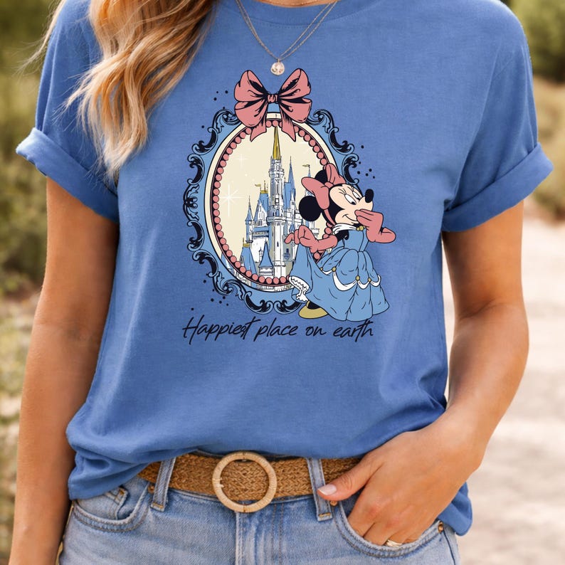 May include: A light blue t-shirt featuring a graphic of Minnie Mouse in a blue dress, standing in front of a castle. The graphic is framed by a pink bow and a decorative oval border. The text "Happiest place on earth" is below the graphic.