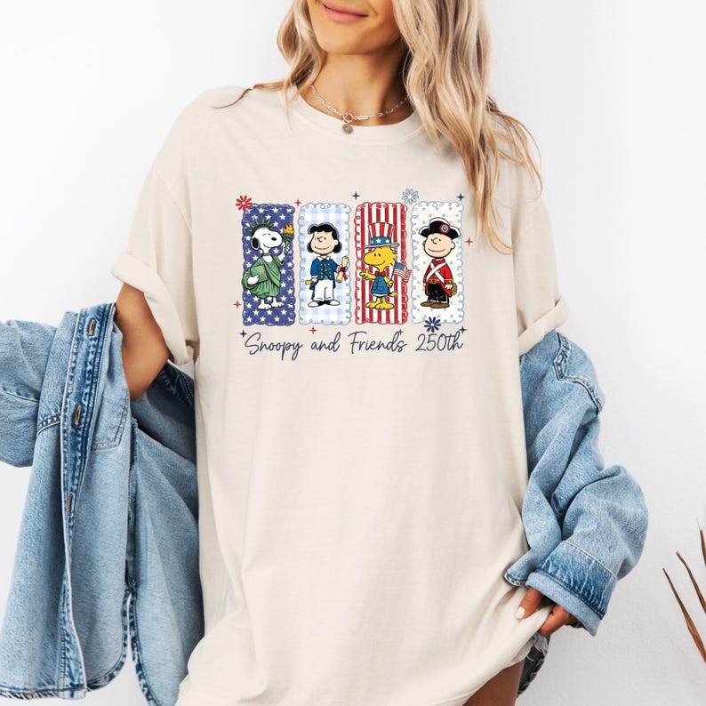 Puede incluir: Camiseta color crema con Snoopy y sus amigos con atuendos patri&oacute;ticos. El dise&ntilde;o incluye a Snoopy, Charlie Brown, Lucy y Linus en marcos rectangulares con estrellas y rayas. El texto en la parte inferior dice "Snoopy and Friends 250th".
