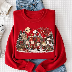 A red sweater with a Christmas-themed design featuring characters from the Peanuts comic strip, including Snoopy, Charlie Brown, and Woodstock, along with a snowy winter scene.