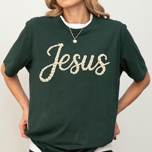 May include: Dark green t-shirt with the word "Jesus" spelled out in a cursive font using off-white pearls. The shirt has short sleeves and a white undershirt is visible at the neckline. A gold necklace with a small pendant is also visible.