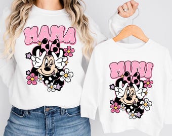 May include: Two white long-sleeve sweatshirts featuring a graphic print of a cartoon mouse with a polka dot bow, making a peace sign. The larger sweatshirt has the text MAMA in pink, and the smaller sweatshirt has the text MINI in pink.