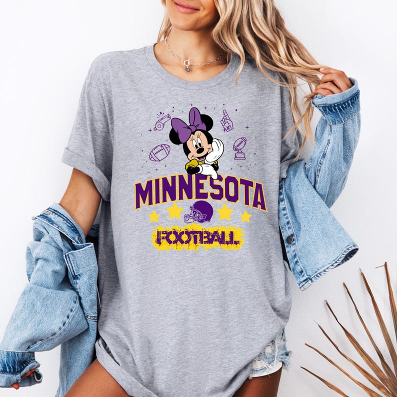 May include: A gray t-shirt featuring a Minnie Mouse design with a purple bow, holding a football. The shirt has the text "MINNESOTA FOOTBALL" in purple and yellow. Additional football-themed graphics are scattered around the design.