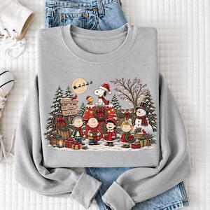 A gray sweatshirt with a Christmas-themed design featuring characters from the Peanuts comic strip, including Snoopy, Woodstock, and other beloved Peanuts characters, along with a snowy winter scene.