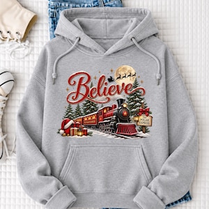 May include: A light gray hooded sweatshirt featuring a graphic of a red steam locomotive train, pine trees, a full moon with a silhouette of a sleigh, and the word Believe in red script. A sign reads The Polar Express near wrapped gifts.