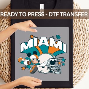 A person is holding a black t-shirt with a graphic design featuring a cartoon character and the word "MIAMI" written on it.