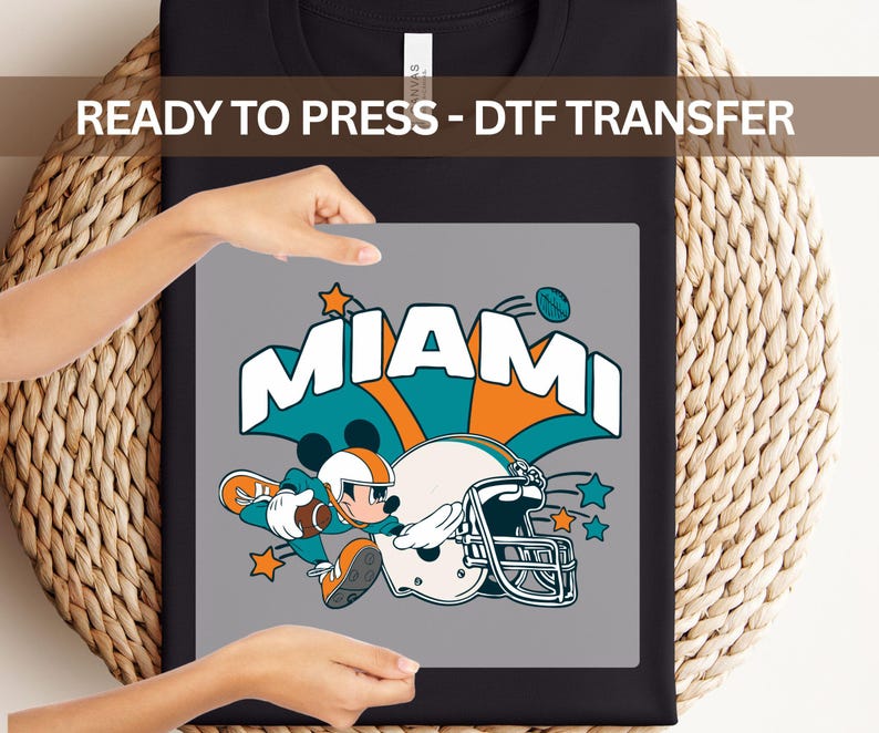 A person is holding a black t-shirt with a graphic design featuring a cartoon character and the word "MIAMI" written on it.
