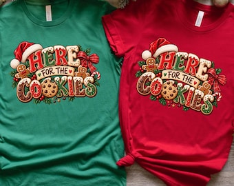 May include: A green t-shirt and a red t-shirt, each featuring a graphic print that reads "HERE FOR THE COOKIES" in gingerbread-style lettering. The design includes a Santa hat, gingerbread people, a red bow, and holly leaves with berries.