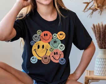 May include: A black short-sleeve t-shirt featuring a graphic print of multiple smiley faces in various sizes and muted colors including yellow, orange, blue, green, and brown. The smiley faces have a distressed, textured appearance.