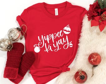 May include: A bright red short-sleeved t-shirt featuring the white text Yippee ki yay in a cursive font, accented with three white Christmas ornament graphics. The shirt is surrounded by two silver ornaments, two red mittens, and red poinsettia flowers.