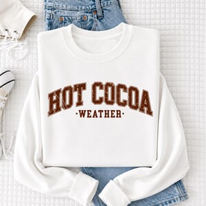 A white sweatshirt with the text "HOT COCOA WEATHER" printed on it, along with a pair of blue jeans and a white sock.