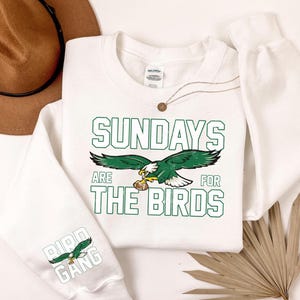 May include: White sweatshirt with the text "SUNDAYS ARE FOR THE BIRDS" in green block letters. A green and white eagle graphic is centered below the text. The sleeve features the text "BIRD GANG" with an eagle graphic.