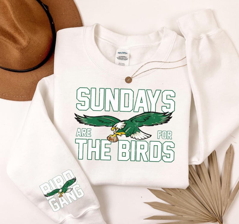 May include: White sweatshirt with the text "SUNDAYS ARE FOR THE BIRDS" in green block letters. A green and white eagle graphic is centered below the text. The sleeve features the text "BIRD GANG" with an eagle graphic.