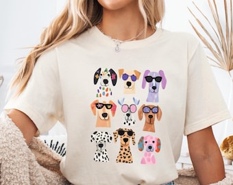 Op de afbeelding: Een crèmekleurig T-shirt met korte mouwen en een grafische print van negen gestileerde hondenkoppen in een raster. Elke hond heeft een ander vachtpatroon en kleur en draagt verschillende soorten kleurrijke zonnebrillen en accessoires.