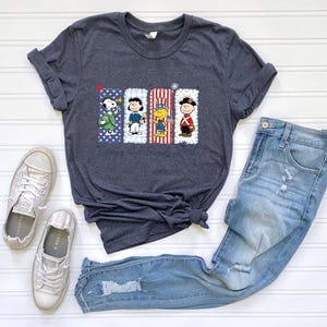 Puede incluir: Camiseta gris oscuro con un gr&aacute;fico de Snoopy y sus amigos con trajes patri&oacute;ticos. El dise&ntilde;o incluye a Snoopy, Charlie Brown, Woodstock y Linus. El texto "Snoopy and Friends 250th" est&aacute; impreso debajo. La camiseta se combina con vaqueros azules y zapatillas.