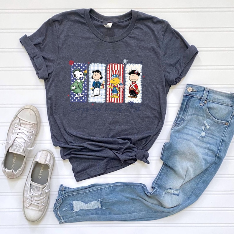 Puede incluir: Camiseta gris oscuro con un gr&aacute;fico de Snoopy y sus amigos con trajes patri&oacute;ticos. El dise&ntilde;o incluye a Snoopy, Charlie Brown, Woodstock y Linus. El texto "Snoopy and Friends 250th" est&aacute; impreso debajo. La camiseta se combina con vaqueros azules y zapatillas.