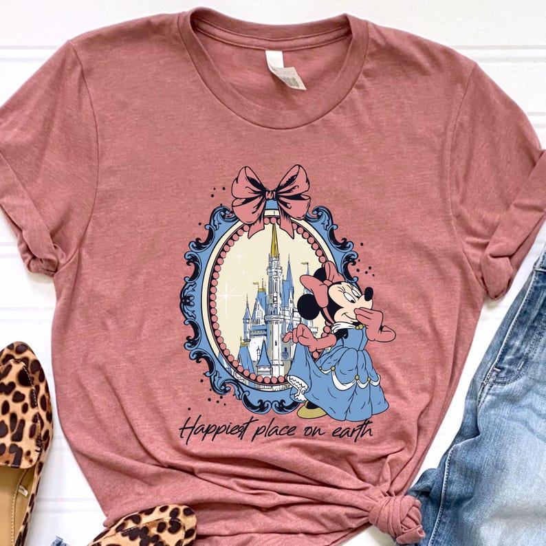 May include: A dusty rose t-shirt featuring a Minnie Mouse graphic in a blue dress, standing in front of a castle. The graphic is framed by a blue and pink oval with a bow at the top. The text "Happiest place on earth" is below the graphic.