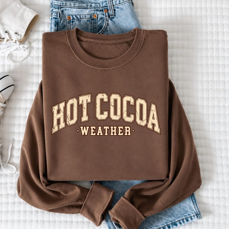 A brown sweatshirt with the text "HOT COCOA WEATHER" printed on it, along with a pair of blue jeans and white sneakers.
