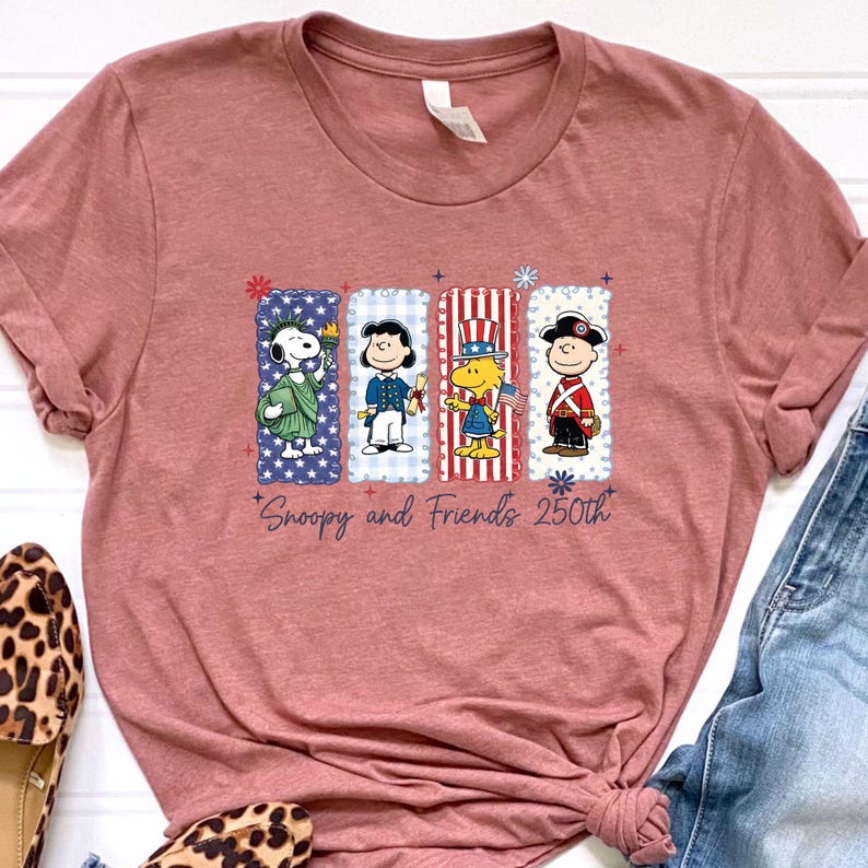 Puede incluir: Camiseta rosa polvoriento con un gr&aacute;fico patri&oacute;tico de Snoopy y sus amigos. El dise&ntilde;o incluye a Snoopy como la Estatua de la Libertad y otros personajes de Peanuts en rojo, blanco y azul. El texto dice "Snoopy and Friends 250th."