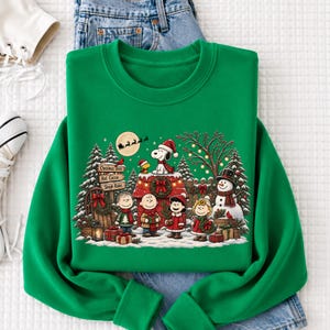 A green sweatshirt with a Christmas-themed design featuring characters from the Peanuts comic strip, including Snoopy, Charlie Brown, and Woodstock, along with a wreath and presents.
