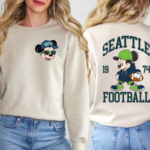 Puede incluir: Sudadera beige con un rat&oacute;n de dibujos animados con gafas de sol y una gorra de b&eacute;isbol con "Seattle". La parte posterior de la sudadera tiene un dise&ntilde;o a juego con la palabra "SEATTLE", los n&uacute;meros "19" y "74" y la palabra "FOOTBALL".