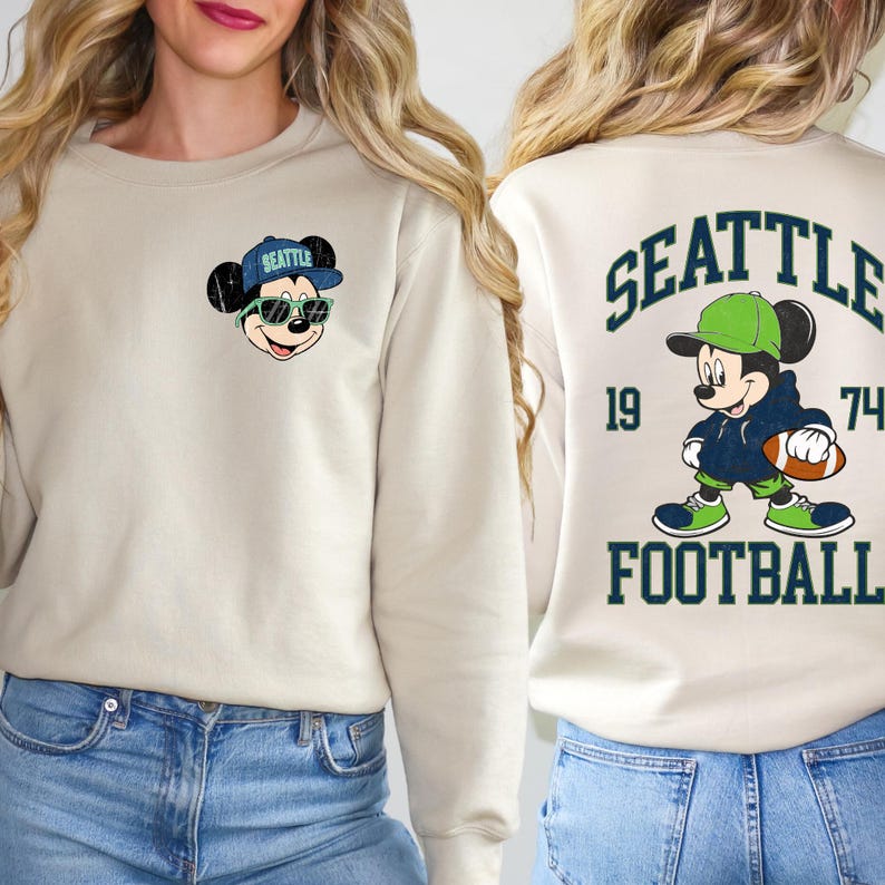 Puede incluir: Sudadera beige con un rat&oacute;n de dibujos animados con gafas de sol y una gorra de b&eacute;isbol con "Seattle". La parte posterior de la sudadera tiene un dise&ntilde;o a juego con la palabra "SEATTLE", los n&uacute;meros "19" y "74" y la palabra "FOOTBALL".