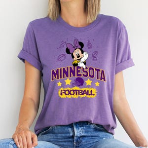May include: A purple t-shirt featuring a Minnie Mouse graphic with a football theme. The shirt has the word "MINNESOTA" in gold lettering, along with stars, a football helmet, and the word "FOOTBALL".