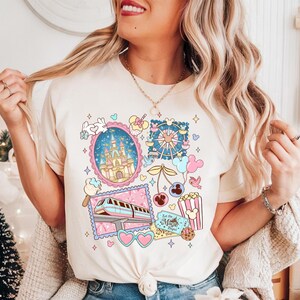 May include: Cream-colored t-shirt featuring a colorful graphic with a castle, Ferris wheel, train, and other whimsical elements. The design includes the text "Let the Magic Begin" and various heart and Mickey Mouse-shaped details.