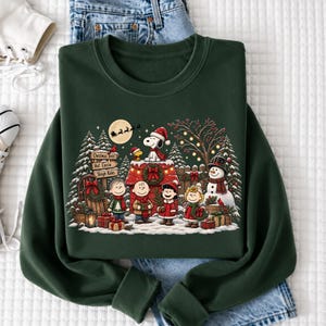 A green sweatshirt with a Christmas-themed design featuring characters from the Peanuts comic strip, including Snoopy, Charlie Brown, and their friends, surrounded by holiday decorations and a snowy landscape.